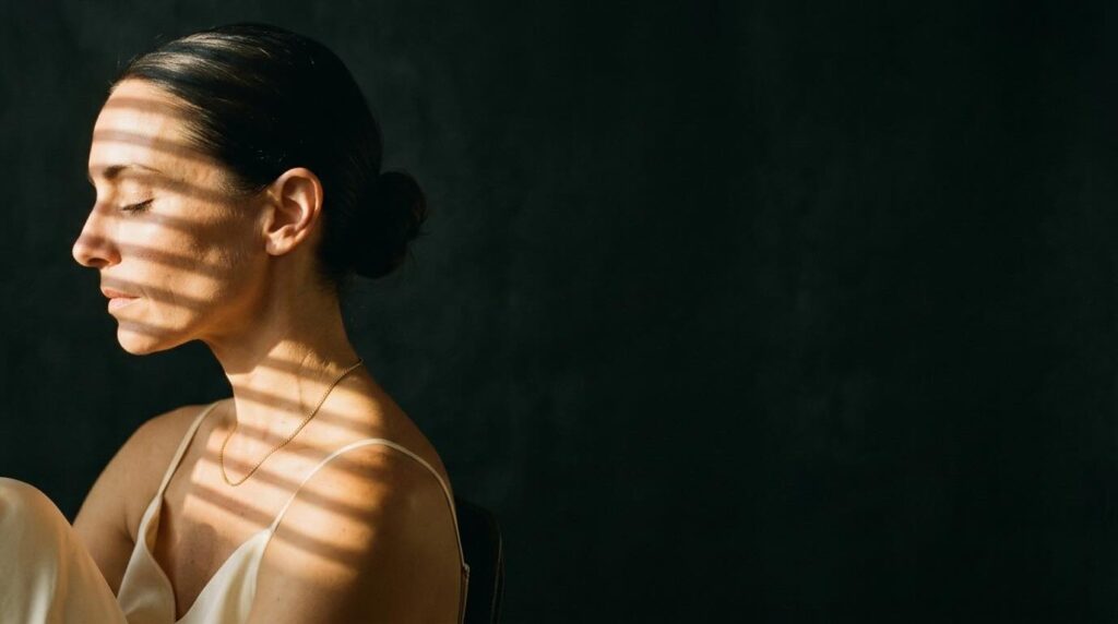 High-contrast editorial side-profile portrait of a woman in her early 40s in pure profile against a deep charcoal background, with warm golden afternoon light filtering through Venetian blinds casting clean horizontal stripes across her cheekbone and shoulder