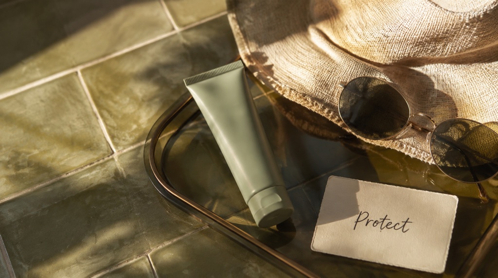 An unbranded sage tube, sun hat and sunglasses beside a handwritten Protect card on olive tiles