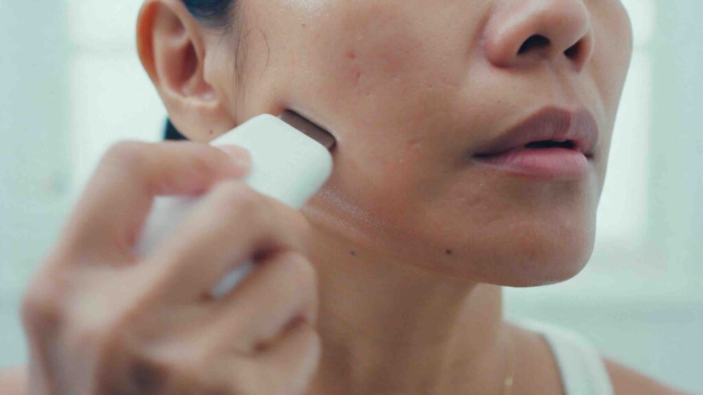 Tight editorial crop of the lower half of a Filipino-American woman's face as she glides a slim white handheld device upward along her jawline in cool daylight her hand softly blurred mid-glide