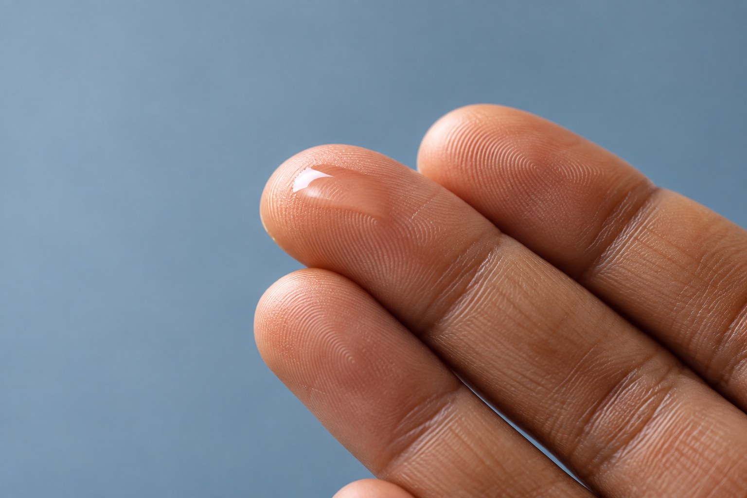 A small clear liquid drop rests on an adult fingertip against a blue-grey background.