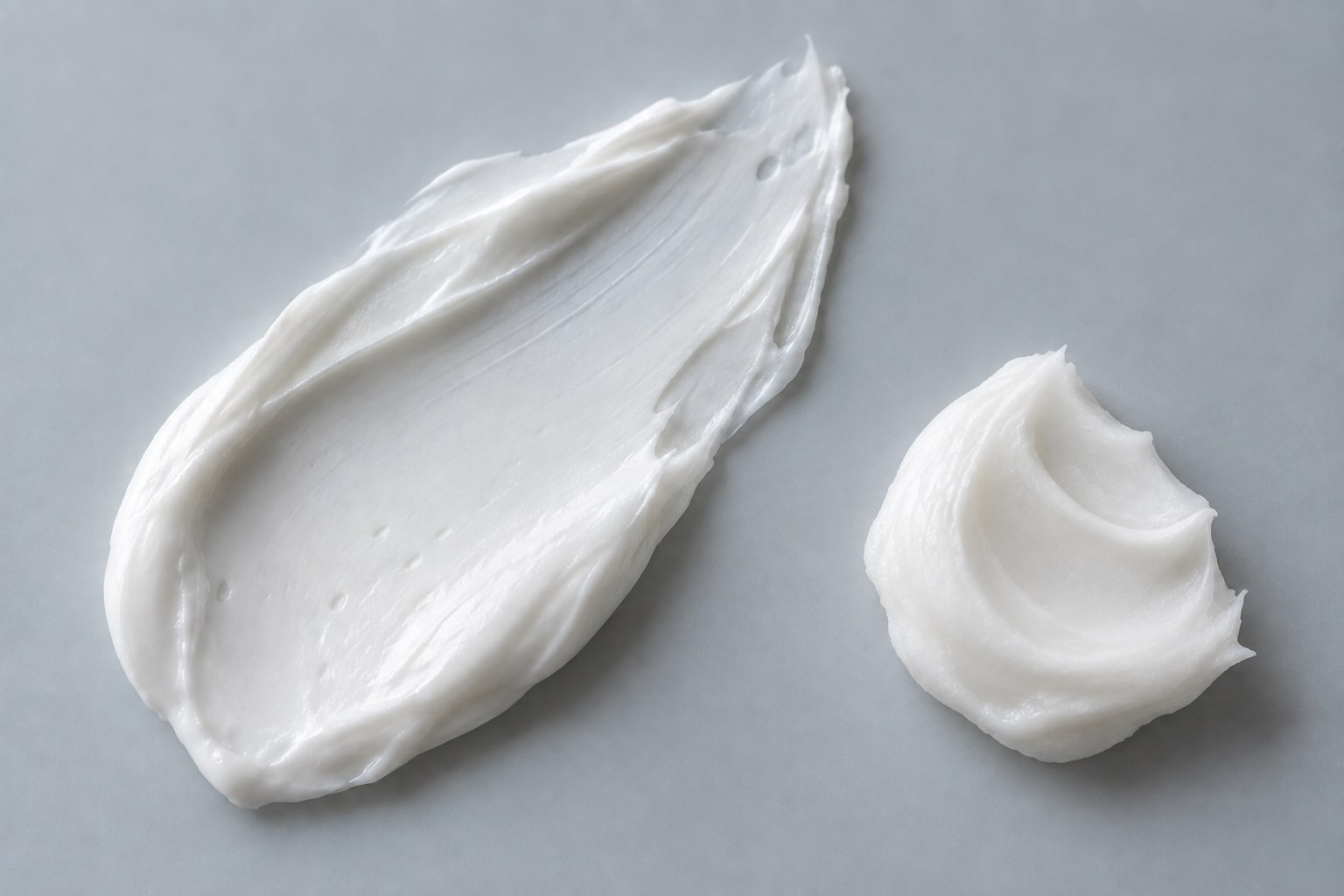 Broad white cream swatch beside a smaller smooth white balm dab on grey glass.