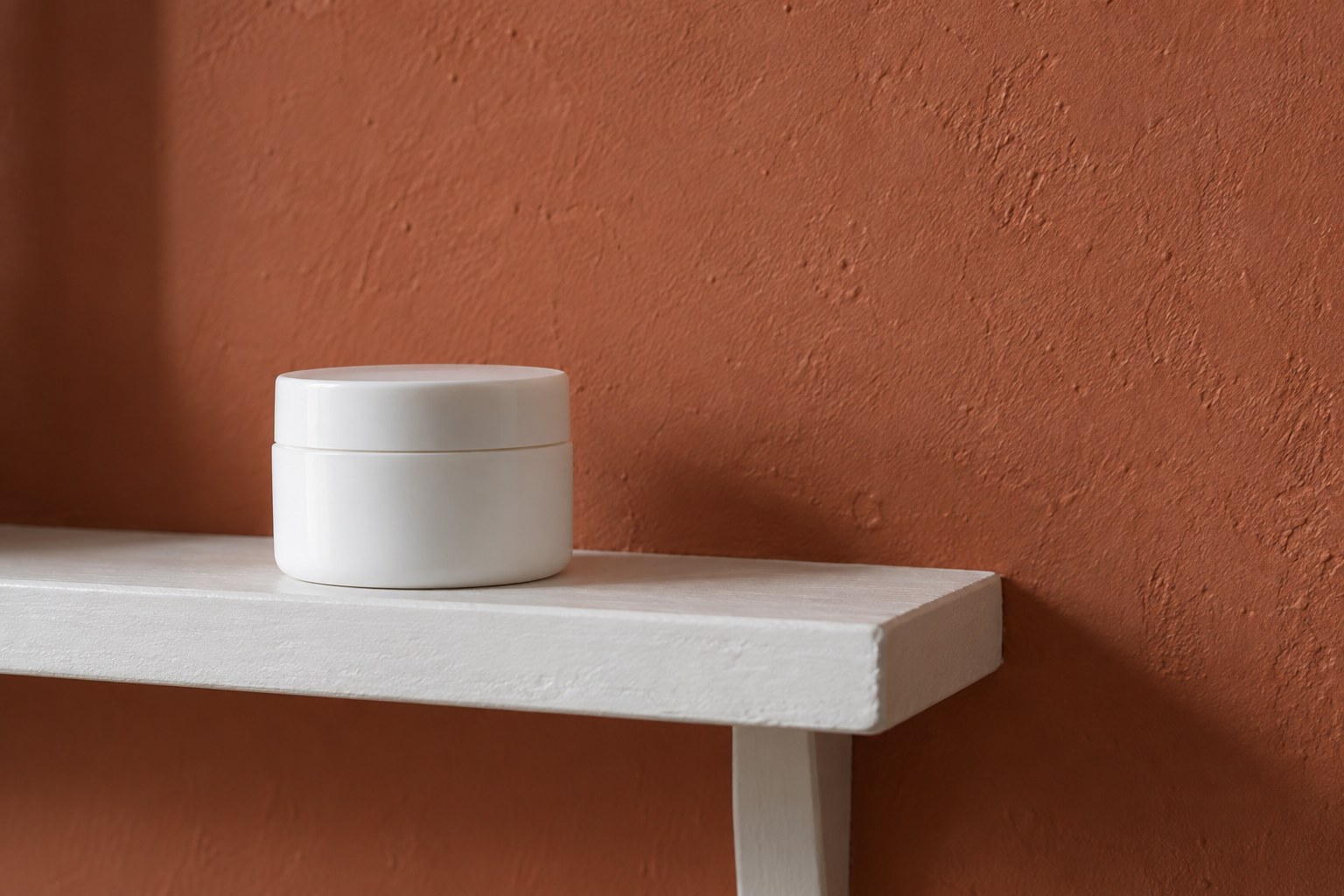 One plain white moisturiser jar on a white shelf against a terracotta wall.