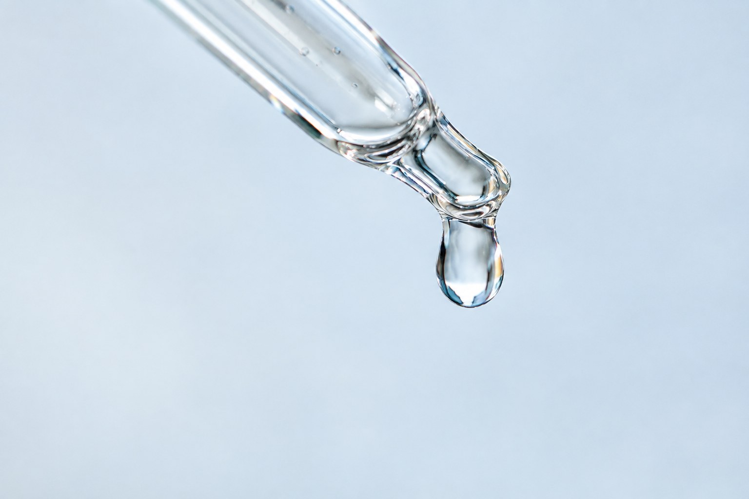 Clear droplet suspended from the tip of a glass pipette against a pale blue background.
