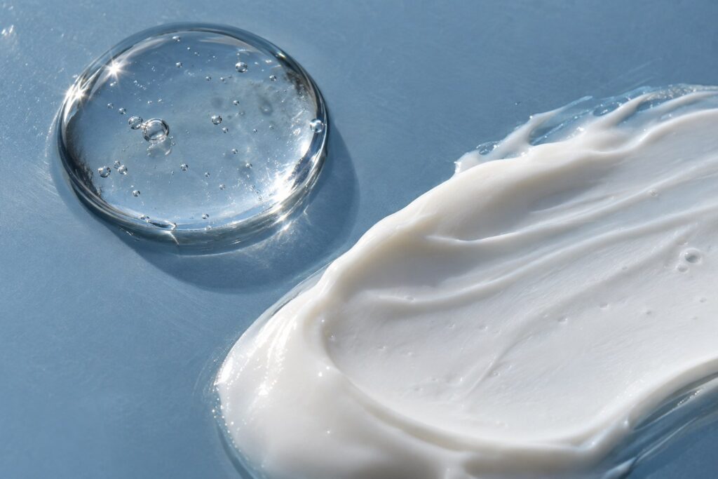 Clear serum droplet beside a textured white moisturiser smear on slate-blue glass.