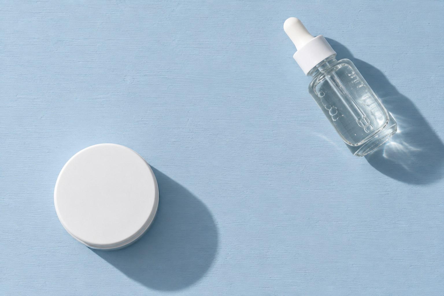 Overhead view of an unlabelled white moisturiser jar and a separate clear serum bottle on a pale blue shelf surface.